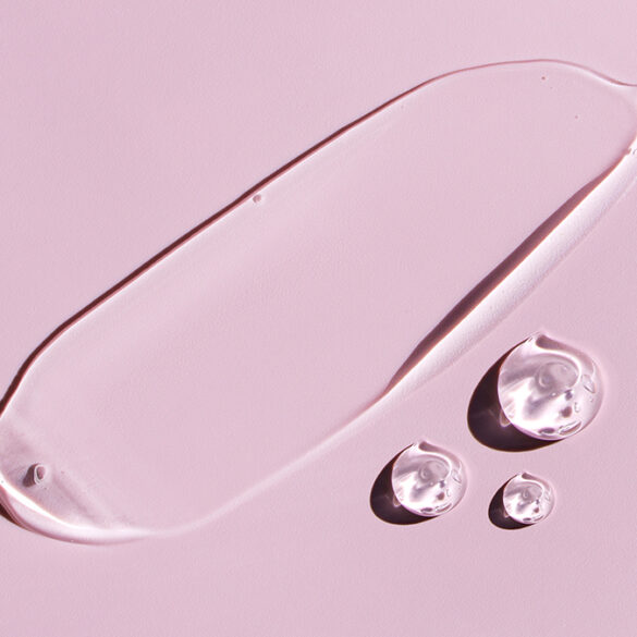 Transparent cosmetic gel or serum smeared over a pink surface alongside three gel drops
